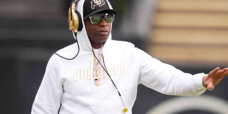 Deion Sanders, head coach of the Colorado Buffaloes, in a white hoodie, hat, sunglasses, and gold headphones, on the field.