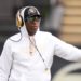Deion Sanders, head coach of the Colorado Buffaloes, in a white hoodie, hat, sunglasses, and gold headphones, on the field.