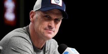 Dan Hurley speaks during a news conference, wearing a blue and white UConn Huskies baseball cap and a grey t-shirt.
