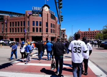 People walking towards Coors Field, a large brick baseball stadium, on a sunny day.