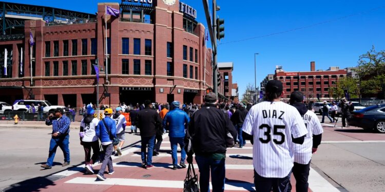 People walking towards Coors Field, a large brick baseball stadium, on a sunny day.