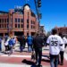 People walking towards Coors Field, a large brick baseball stadium, on a sunny day.
