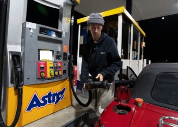 A person in a gray beanie and a black hoodie filling the tank of their red Mazda Miata at an Astro gas station.