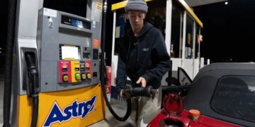 A person in a gray beanie and a black hoodie filling the tank of their red Mazda Miata at an Astro gas station.