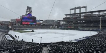 Snow-covered baseball field with visible stadium seats and large screens under a cloudy sky.