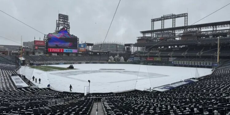 Snow-covered baseball field with visible stadium seats and large screens under a cloudy sky.