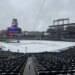 Snow-covered baseball field with visible stadium seats and large screens under a cloudy sky.