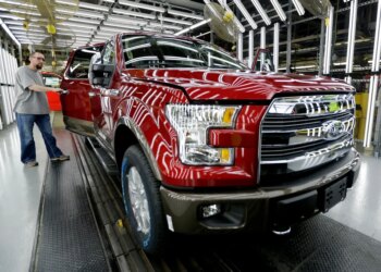A worker inspects a red Ford F-150 truck with an open door at an assembly plant.