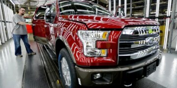 A worker inspects a red Ford F-150 truck with an open door at an assembly plant.