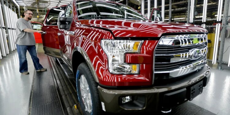 A worker inspects a red Ford F-150 truck with an open door at an assembly plant.