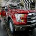 A worker inspects a red Ford F-150 truck with an open door at an assembly plant.