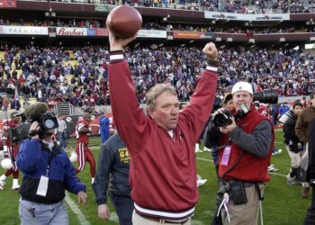 Arizona Cardinals head coach Dave McGinnis celebrates a win, raising a football in one hand and pumping his other fist in the air, on a football field filled with players and media.