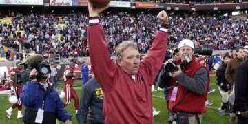 Arizona Cardinals head coach Dave McGinnis celebrates a win, raising a football in one hand and pumping his other fist in the air, on a football field filled with players and media.
