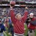 Arizona Cardinals head coach Dave McGinnis celebrates a win, raising a football in one hand and pumping his other fist in the air, on a football field filled with players and media.