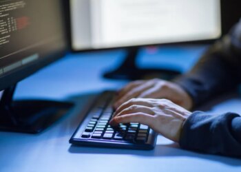 Hacker's hands typing on a keyboard in front of a monitor displaying code.