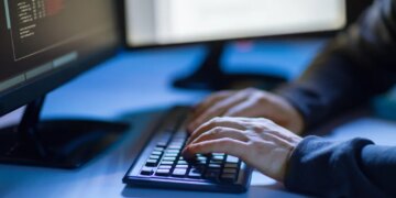 Hacker's hands typing on a keyboard in front of a monitor displaying code.
