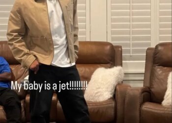 Kenyon Sadiq in a tan jacket, white shirt, and black pants, with a green and white cap on his head, standing on a rug in front of two brown leather recliners with white pillows.