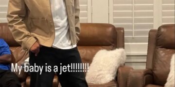 Kenyon Sadiq in a tan jacket, white shirt, and black pants, with a green and white cap on his head, standing on a rug in front of two brown leather recliners with white pillows.