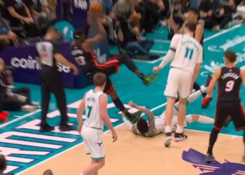 A basketball player in a black and red uniform holding a basketball, jumping over another player in a white and green uniform who has fallen to the ground.