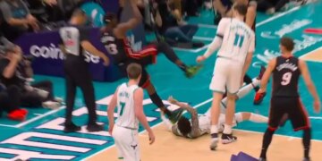 A basketball player in a black and red uniform holding a basketball, jumping over another player in a white and green uniform who has fallen to the ground.