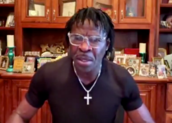 Deion Sanders wearing glasses and a black shirt with a silver cross necklace.