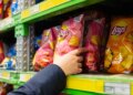 A person's hand reaching for a pink bag of Lay's potato chips on a store shelf.