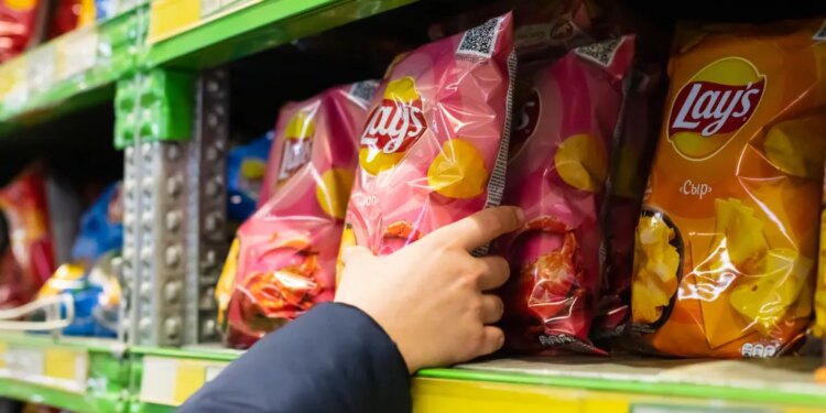 A person's hand reaching for a pink bag of Lay's potato chips on a store shelf.