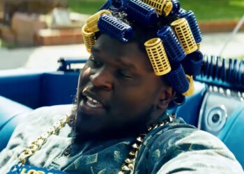 Male in a blue car wearing blue and yellow hair curlers and a gold chain.