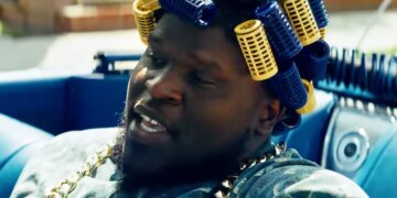 Male in a blue car wearing blue and yellow hair curlers and a gold chain.