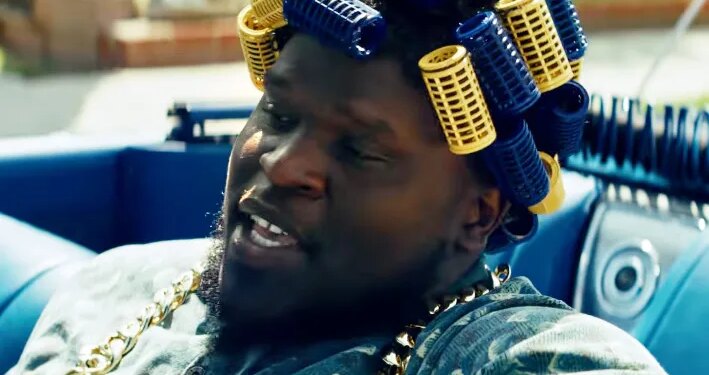 Male in a blue car wearing blue and yellow hair curlers and a gold chain.