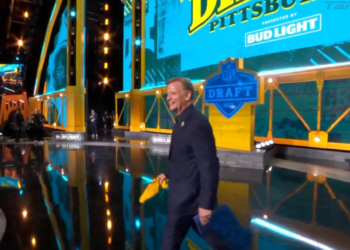 A man in a suit carrying papers walks across a stage for the NFL Draft presented by Bud Light.