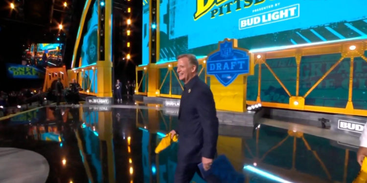 A man in a suit carrying papers walks across a stage for the NFL Draft presented by Bud Light.