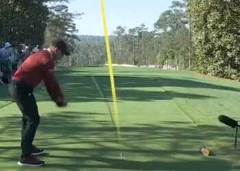 A golfer in a red shirt and black pants mid-swing on a golf course.
