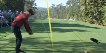 A golfer in a red shirt and black pants mid-swing on a golf course.