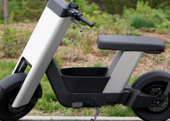A photo of a silver e-bike from the side, in front of trees.
