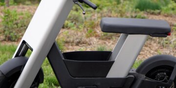 A photo of a silver e-bike from the side, in front of trees.