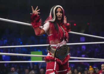 Mercedes Mone stands in a wrestling ring, wearing a red and white outfit with fuzzy armbands and a championship belt around her waist.