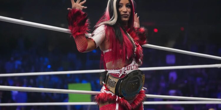 Mercedes Mone stands in a wrestling ring, wearing a red and white outfit with fuzzy armbands and a championship belt around her waist.
