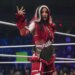Mercedes Mone stands in a wrestling ring, wearing a red and white outfit with fuzzy armbands and a championship belt around her waist.
