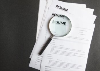 A magnifying glass rests on a stack of resumes on a black surface.