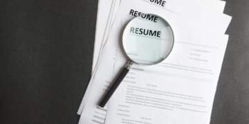A magnifying glass rests on a stack of resumes on a black surface.