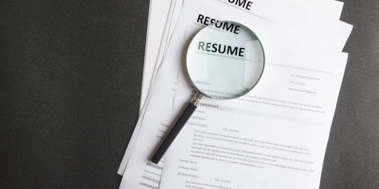 A magnifying glass rests on a stack of resumes on a black surface.