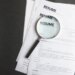 A magnifying glass rests on a stack of resumes on a black surface.