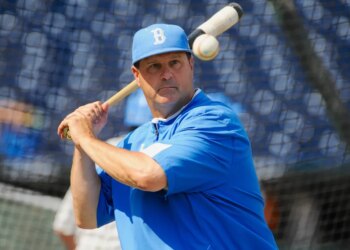 A baseball player in a blue cap and shirt holds a bat with a baseball above his shoulder.