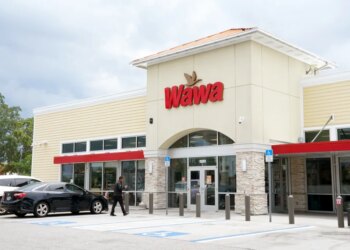 Wawa convenience store exterior with a red and brown logo, a man walking on the sidewalk, and cars parked outside.
