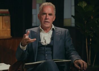 Jordan Peterson talking and gesturing with his right hand while wearing a dark suit.