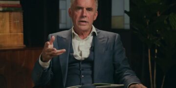 Jordan Peterson talking and gesturing with his right hand while wearing a dark suit.