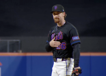 A baseball player in a black Mets uniform with the number 26 and a New York-Presbyterian patch on his sleeve, stands with his hand over his heart.