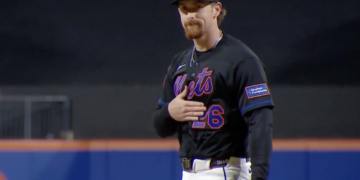 A baseball player in a black Mets uniform with the number 26 and a New York-Presbyterian patch on his sleeve, stands with his hand over his heart.