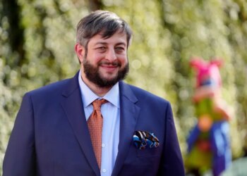 Eugene Mirman, wearing a blue suit, orange tie, and pocket square, smiles at the camera.